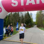 Młody biegacz przekracza linię startową pod różowo-niebieskim balonem. W tle widoczny las i grupa dzieci kibicujących. Pogoda jest pochmurna.