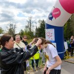 Kobieta w ciemnej kurtce wręcza medal uczestniczce w białej koszulce. W tle niebiesko-biały balon i grupa osób. Scena na świeżym powietrzu z zielenią i pochmurnym niebem.