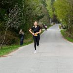 Biegaczka w czarnym stroju biegnie po wiejskiej drodze otoczonej drzewami. W tle inni uczestnicy i widzowie. Zgaszone kolory natury.