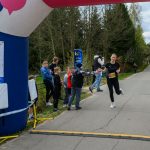 Biegaczka na mecie pod granatowym łukiem na szarej drodze. Z boku dzieci i drzewa. Kolory: zielony, granatowy.