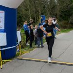 Na obrazku kobieta w czarnym stroju sportowym biegnie przez linię mety. W tle widać kibiców i zielone drzewa. Po lewej stronie znajduje się niebieski balon startowy z mapą.