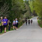 Grupa biegaczy na asfaltowej drodze w lesie. Ubrani w kolorowe stroje z numerami startowymi. Tło zielone, otoczone drzewami.