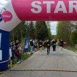 Początek biegu pod różowym balonem z napisem START. Biegacze w czarnych strojach trzymają się za ręce. W tle drzewa i kibice na poboczu.