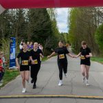 Na zdjęciu cztery biegaczki przekraczają linię mety, trzymając się za ręce. Ubrane na czarno, na tle zielonych drzew. Dookoła widzowie i niebiesko-różowy baner.