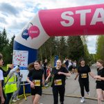Biegacze w czerni przekraczają linię startu. Balon startowy w kolorach różowym i białym. Tło to drzewa i niebo z chmurami.