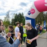 Uczestnicy biegu przekraczają różowy łuk mety. Mężczyzna wręcza medale. W tle drzewa i chmury.