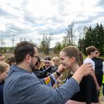 Mężczyzna wręcza medal uśmiechniętej dziewczynie. W tle ludzie, drzewa i pochmurne niebo. Z prawej widoczny balon reklamowy.