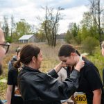 Na zdjęciu osoba zakłada medal na szyję uczestniczki biegu. W tle zielone krzewy i dom przy pochmurnym niebie. Ubrani są w czarne koszulki.