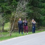 Trzy osoby stoją obok drogi w lesie, na tle zielonych drzew. Jedna z nich trzyma telefon w rękach. Scena jest wiosenna.