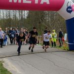 Trzech młodych biegaczy startuje spod różowo-niebieskiego, dmuchanego łuku z napisem META. W tle widoczny jest tłum ludzi stojący na poboczu wśród drzew. Obok ustawiony jest elektroniczny zegar pomiarowy, wyświetlający czas