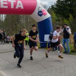 Trzech mężczyzn biegnie przez metę. Nad nimi różowy łuk z napisem META. W tle widoczni kibice i drzewa.