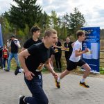 Na zdjęciu widać trzech biegnących młodych ludzi w czarno-białych strojach. W tle znajduje się las oraz niebieski baner reklamowy. Po lewej stronie widać nadmuchiwany łuk startowy w biało-różowych barwach.