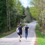 Na zdjęciu trzech biegaczy na wiejskiej drodze. Otoczenie to zielony las i drzewa. W tle widoczni są widzowie przy drodze.