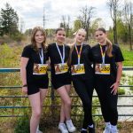 Cztery osoby w czarnych strojach stoją na tle przyrody z medalami na szyjach. Mają numery startowe. Wiosenne drzewa w tle.