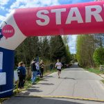 Biegacz przekracza linię startu pod różowo-białym łukiem z napisem START. W tle zielone drzewa i niebieskie niebo. Kibice stoją po bokach drogi.