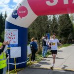 Zawodnik w białej koszulce przekracza linię mety pod dmuchanym łukiem. Tło to droga otoczona drzewami. Obok ludzie kibicują.