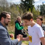 Na zdjęciu widać ludzi na świeżym powietrzu klaskających sobie w dłonie. Młodzieniec w białej koszulce ma żółty numer startowy. W tle są drzewa i zarośla, a niebo jest częściowo zachmurzone.