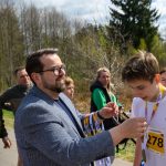 Mężczyzna w okularach wręcza medal młodemu biegaczowi w białej koszulce. W tle znajduje się grupa osób oraz las. Dominują odcienie zieleni i szarości.