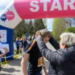 Na zdjęciu osoba wręcza medal biegaczowi pod różowym łukiem z napisem START. W tle grupa ludzi i drzewa. Słońce świeci, tworząc jasną atmosferę.