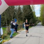 Biegacz w czarnym stroju kończy bieg pod różowo-niebieskim balonem. Droga otoczona zielonym lasem. Na boku stoi fotograf.