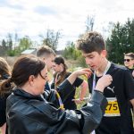 Grupa ludzi na tle drzew. Kobieta wręcza medale uczestnikom biegu w czarnych koszulkach. Żółte numery startowe.