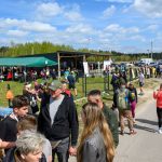 Grupa ludzi na świeżym powietrzu podczas wydarzenia na tle zieleni i namiotów. Niebo jest niebieskie, pełne chmur. Uczestnicy są ubrani kolorowo, w tle widać samochody.