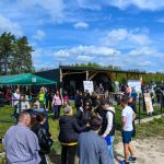 Na zdjęciu festyn plenerowy z grupą ludzi. Namiot w kolorze zielonym, na tle niebieskiego nieba z chmurami. W tle las i budynek.