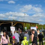 Tłum ludzi przed drewnianym budynkiem na tle zielonych drzew. Niebieskie, pochmurne niebo. Scena z transparentami i plakatami.