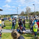 Na zdjęciu jest tłum ludzi na zewnątrz w słoneczny dzień. Dominują kolory zielony, niebieski i różne odcienie ubrań. W tle widać samochody i drzewa.