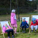 Dzieci siedzą na leżakach na zielonej trawie. Jedno dziecko w różowym bezrękawniku stoi, inne w czerwonych i niebieskich kurtkach siedzą. Tło to ogrodzenie i drzewa.