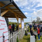 Na zdjęciu grupka ludzi na zewnątrz przy drewnianym zadaszeniu. Tło to błękitne niebo z chmurami i drzewa. Widoczne są banery z logo Mazowsze.