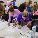 Grupa osób przy stole podpisująca dokumenty. Tło jasne, dominują kolory fioletowy, różowy oraz czarny. Na stole butelki z wodą.