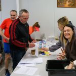 Grupa ludzi uczestniczy w rejestracji w pomieszczeniu z białymi ścianami. Na stole są papiery i butelki wody. Kobieta w brunatnej bluzie uśmiecha się i pokazuje znak pokoju.