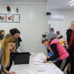 Na obrazie grupa ludzi w pomieszczeniu podpisuje dokumenty przy białym stole. Ściany są białe, z rysunkami zwierząt. W tle widać różnorodne osoby, niektóre ubrane kolorowo.