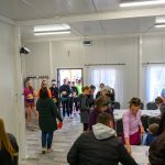 Grupa ludzi w kolorowych sportowych ubraniach czeka w jasnym pomieszczeniu z oknami w tle. Wnętrze ma białe ściany i podłogę w szarych tonach. Na stole przed nimi leżą dokumenty.