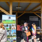 Na zdjęciu widoczny jest drewniany budynek z tablicą Świetlica Wólka Kokosia. Ludzie stoją przy dwóch planszach promocyjnych Gminy Dobre. Jasne kolory ubrań kontrastują z drewnianą konstrukcją.