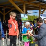 Na zdjęciu są trzy dzieci z medalami, ubrane kolorowo, odbierające nagrody. W tle widoczna jest drewniana konstrukcja i baner. Tło jest zielone z niebieskim niebem, a kompozycja jest żywa i radosna.