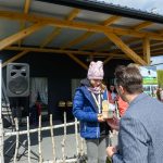 Na zdjęciu jest osoba w granatowej kurtce, która otrzymuje trofeum na zewnątrz. W tle są drewniana wiata i baner z napisem „Mazowsze”. Obok stoją osoby, a pogoda jest słoneczna.