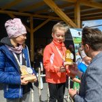 Dwie dziewczynki otrzymują trofea na podium, ubrane w kolorowe kurtki. W tle drewniana altana i ludzie. Słoneczny dzień.