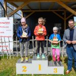 Trzy dziewczynki na podium z trofeami na tle drewnianego budynku. Niebieskie niebo, plakaty i zielona trawa w tle. Obok stoi mężczyzna.