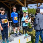Trzech chłopców stoi na podium w niebieskich strojach sportowych. Otrzymują medale i trofea. W tle jest drewniana pergola i zieleń.