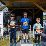 Trzech chłopców na podium z pucharami w rękach. Tło z logiem Mazowsze i drewnianym zadaszeniem. Mężczyzna w marynarce wręcza nagrody.