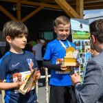 Dwoje dzieci w niebieskich koszulkach trzyma trofea. W tle jasne drewniane zadaszenie i reklama z zielonym krajobrazem. Mężczyzna w okularach rozmawia z jednym z chłopców.