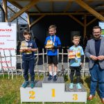 Chłopcy stoją na podium z trofeami. Obok nich mężczyzna w marynarce. Tło to drewniana konstrukcja i plakaty reklamowe.