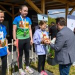 Na zdjęciu widnieją trzy młode dziewczyny na podium w sportowych strojach. Każda ma medal, a jedna z nich trzyma trofeum. W tle widać drewnianą konstrukcję i niebo z chmurami.