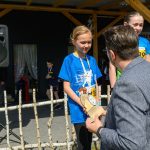 Dziecko w niebieskiej koszulce otrzymuje nagrodę od mężczyzny w marynarce. W tle drewniana konstrukcja i inni uczestnicy.