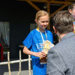 Na zdjęciu dziecko w niebieskiej koszulce otrzymuje trofeum. W tle drewniana konstrukcja i głośnik. Dzień jest słoneczny.