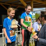 Dwie młode dziewczyny na podium z pucharami, ubrani w kolorowe, sportowe koszulki. Obok nich stoi mężczyzna w marynarce, który wręcza nagrody. Tło z drewnianą konstrukcją i plakatami.