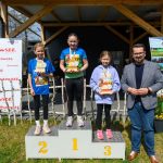 Trzy dziewczynki stoją na podium z trofeami, obok mężczyzna. Tło to drewniana konstrukcja i banery. Kolory jasne, wiosenne.