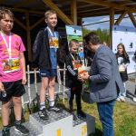 Trzech chłopców stoi na podium. Jeden mężczyzna wręcza im nagrody. W tle drewniana konstrukcja i baner.
